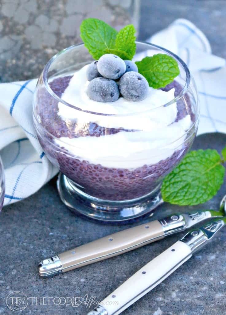 blueberry chia seeds pudding in a clear glass bowl with utensils and green accent mint leaves